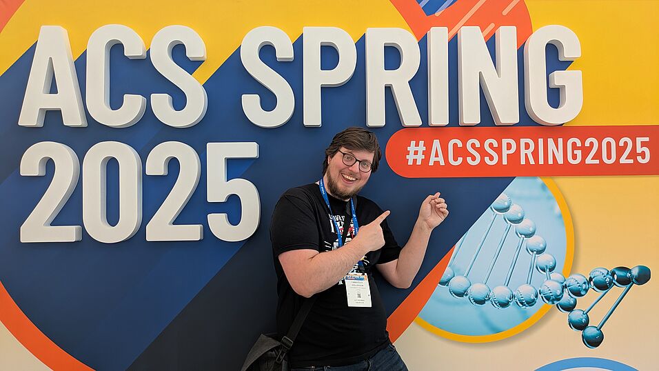 A young man with glasses posing in front of a big "ACS Spring 2025" sign