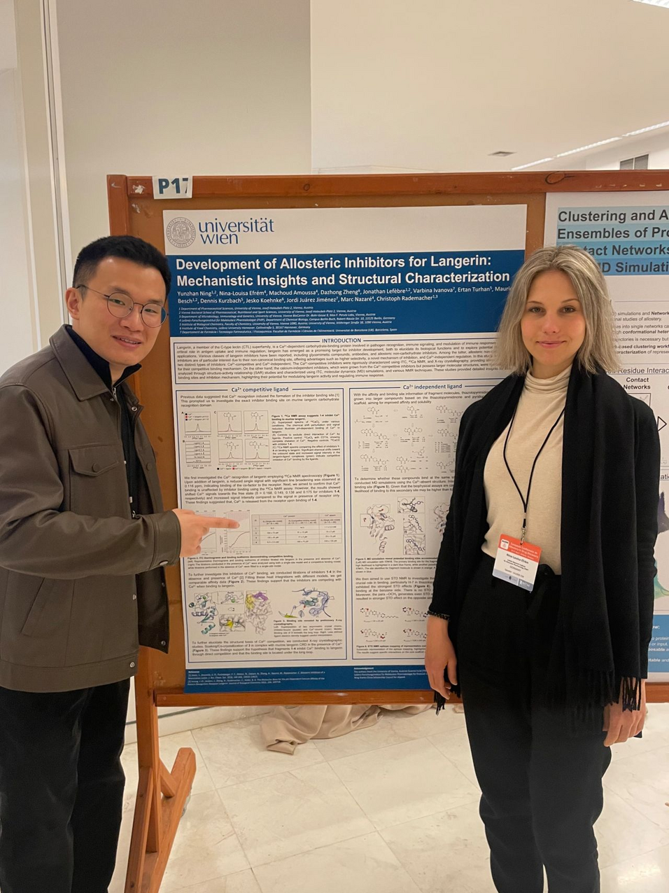 a dark haired man with glasses and a blond girl standing in front of the poster